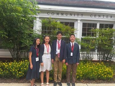 4 young people with medals around their neck standing in front of a white building with green shrubbery in the background