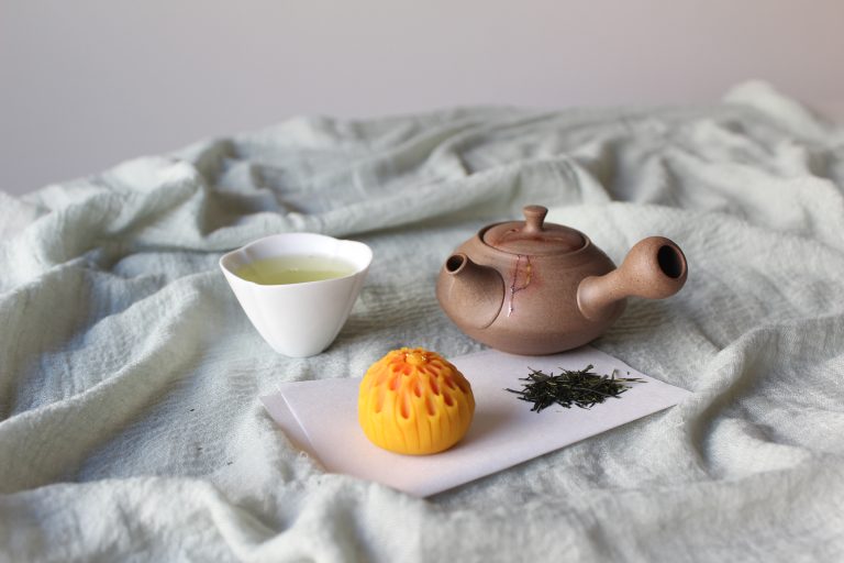 A ceramic teapot, a cup of green tea, a yellow pastry, and loose tea leaves are arranged on a light fabric surface for an inviting Japanese tea tasting experience.