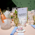 A round table set with vintage teacups, glassware, a teapot, cloth napkins, and a bottle of rosé in ice; a sign reads "Hazeltine's.