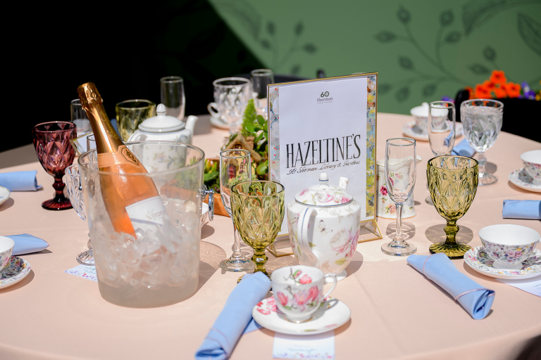 A round table set with vintage teacups, glassware, a teapot, cloth napkins, and a bottle of rosé in ice; a sign reads "Hazeltine's.