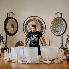 A man in a black t-shirt plays crystal singing bowls in front of three large gongs during a sound bath.