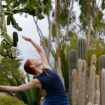A woman with red hair in a ponytail stretches her arms upward, embracing the Garden Flow as she stands among various cacti and tropical plants outdoors.