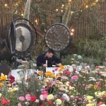 A person sits at a table surrounded by colorful flowers, with two large gongs and string lights in the background, creating a peaceful sound healing atmosphere reminiscent of shinrin-yoku, or forest bathing.