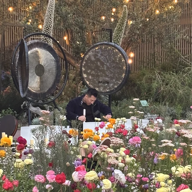 A person sits at a table surrounded by colorful flowers, with two large gongs and string lights in the background, creating a peaceful sound healing atmosphere reminiscent of shinrin-yoku, or forest bathing.