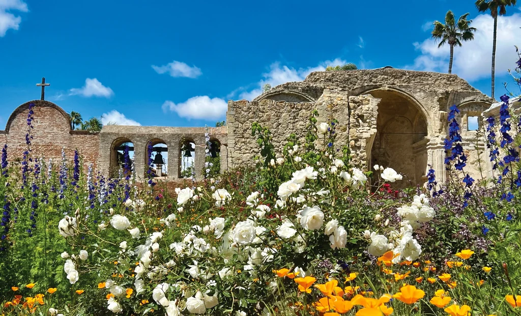 Ruins of an old stone building with arches and a cross at Mission San Juan Capistrano, surrounded by blooming white, orange, and purple flowers under a bright blue sky.