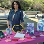 A woman stands behind a table covered with art, chocolates, and a sign reading “There are no mistakes in fluid art, only happy accidents” at an outdoor Acrylic Pour Painting workshop.
