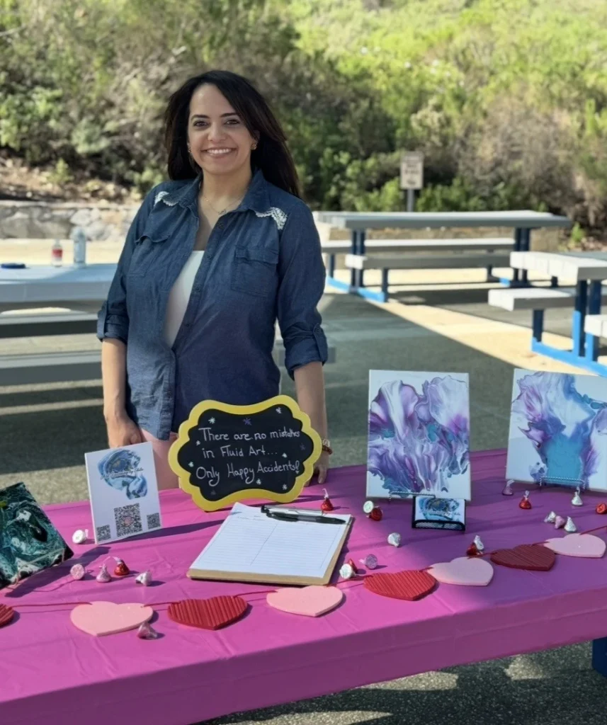 A woman stands behind a table covered with art, chocolates, and a sign reading “There are no mistakes in fluid art, only happy accidents” at an outdoor Acrylic Pour Painting workshop.
