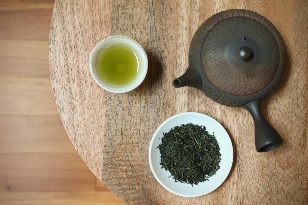 A ceramic teapot, a small cup of Japanese green tea, and a white plate with loose green tea leaves on a wooden table create the perfect setting for an authentic tea tasting experience.