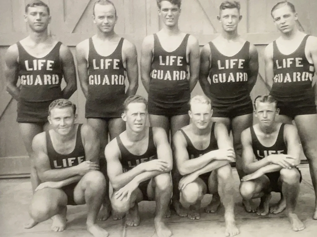 Black-and-white photo of nine men in vintage swimsuits labeled “LIFE GUARD,” posing barefoot in two rows in front of a wooden door, perhaps members of an early lifeguard department captured before tragedy or triumph touched their Russian coastal community.