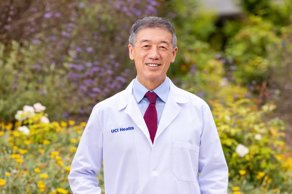 A person in a white UCI Health lab coat and red tie stands outdoors in front of green foliage and yellow flowers, smiling at the camera, embodying a commitment to lifelong health and wellness.