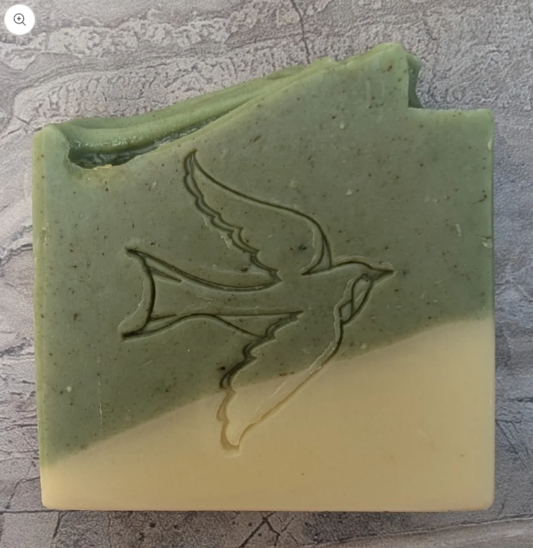 A rectangular, two-toned green and cream natural handmade soap bar with a bird silhouette engraved on the green portion, placed on a textured gray surface.