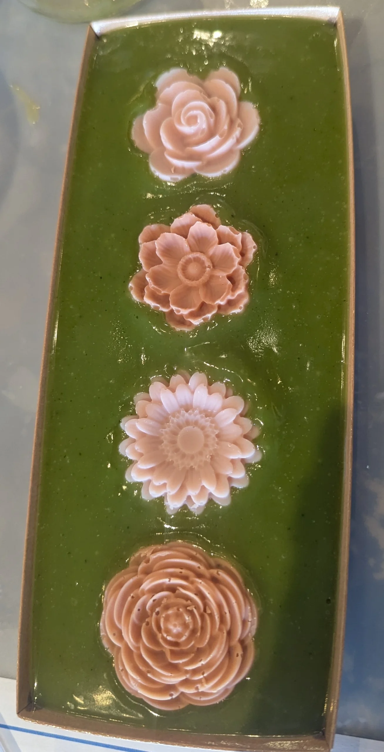 Rectangular tray with green jelly base, topped with three pink, flower-shaped jellies arranged in a vertical line—beautifully reminiscent of handcrafted treats from a natural soap workshop.