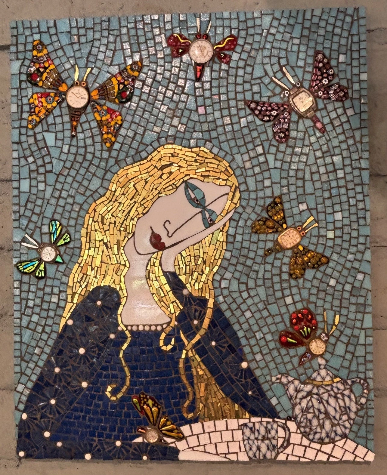 Mosaic artwork of a woman with long yellow hair, blue eyes, and a blue dress sitting at a table with a teapot and cups, surrounded by five colorful butterflies on a blue background, inspired by nature and reminiscent of an Irina Charny art installation.