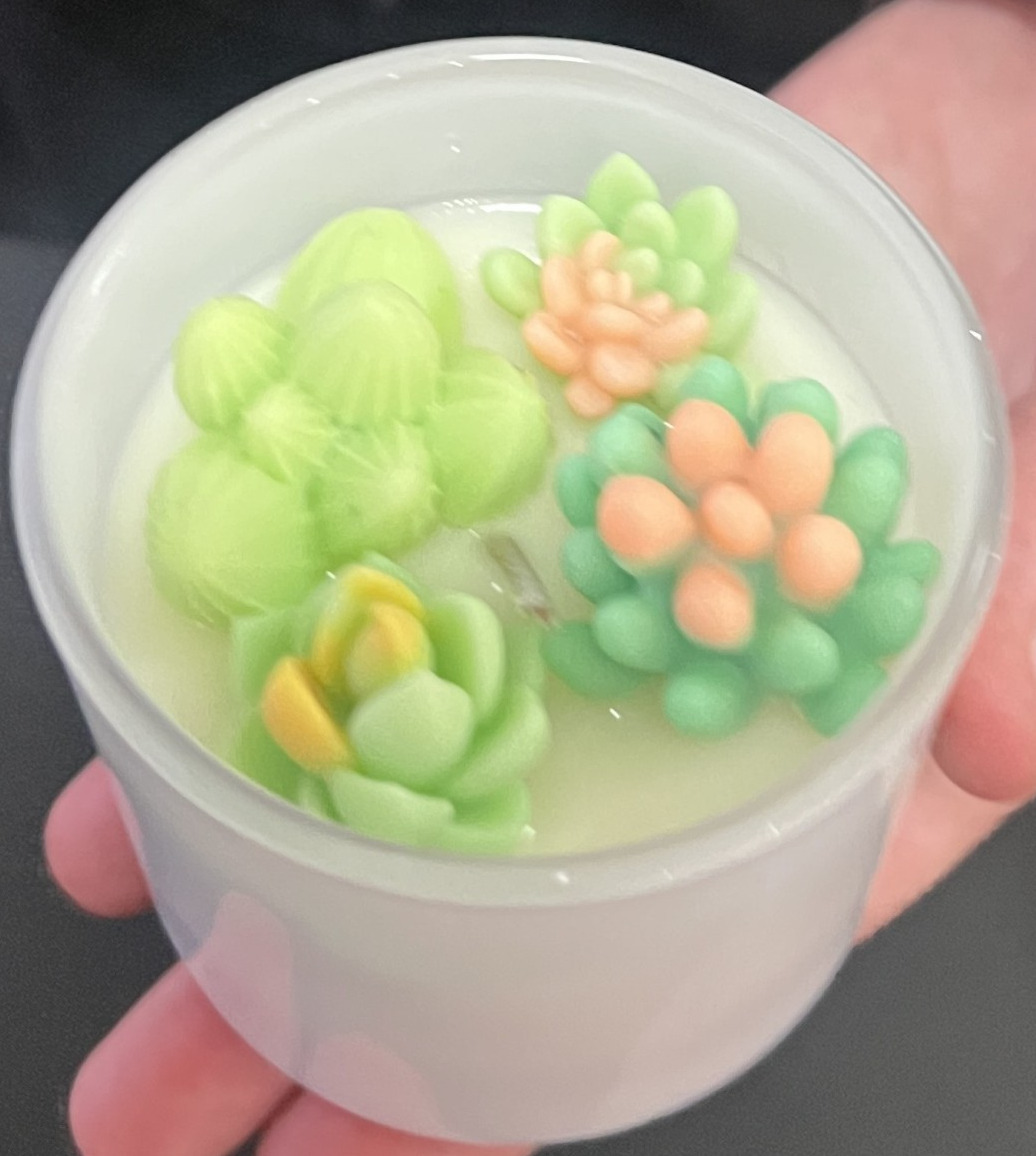 A hand holds a white container with a candle topped with wax decorations shaped like colorful succulents, showcasing the art of botanical candle crafting.