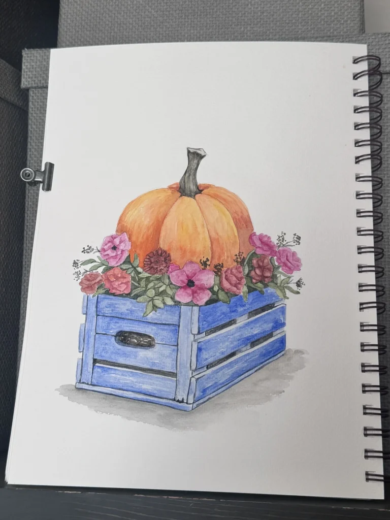 A watercolor drawing of a large pumpkin resting on pink flowers inside a blue crate, sketched on a spiral-bound notebook, captures the gentle charm of autumn.