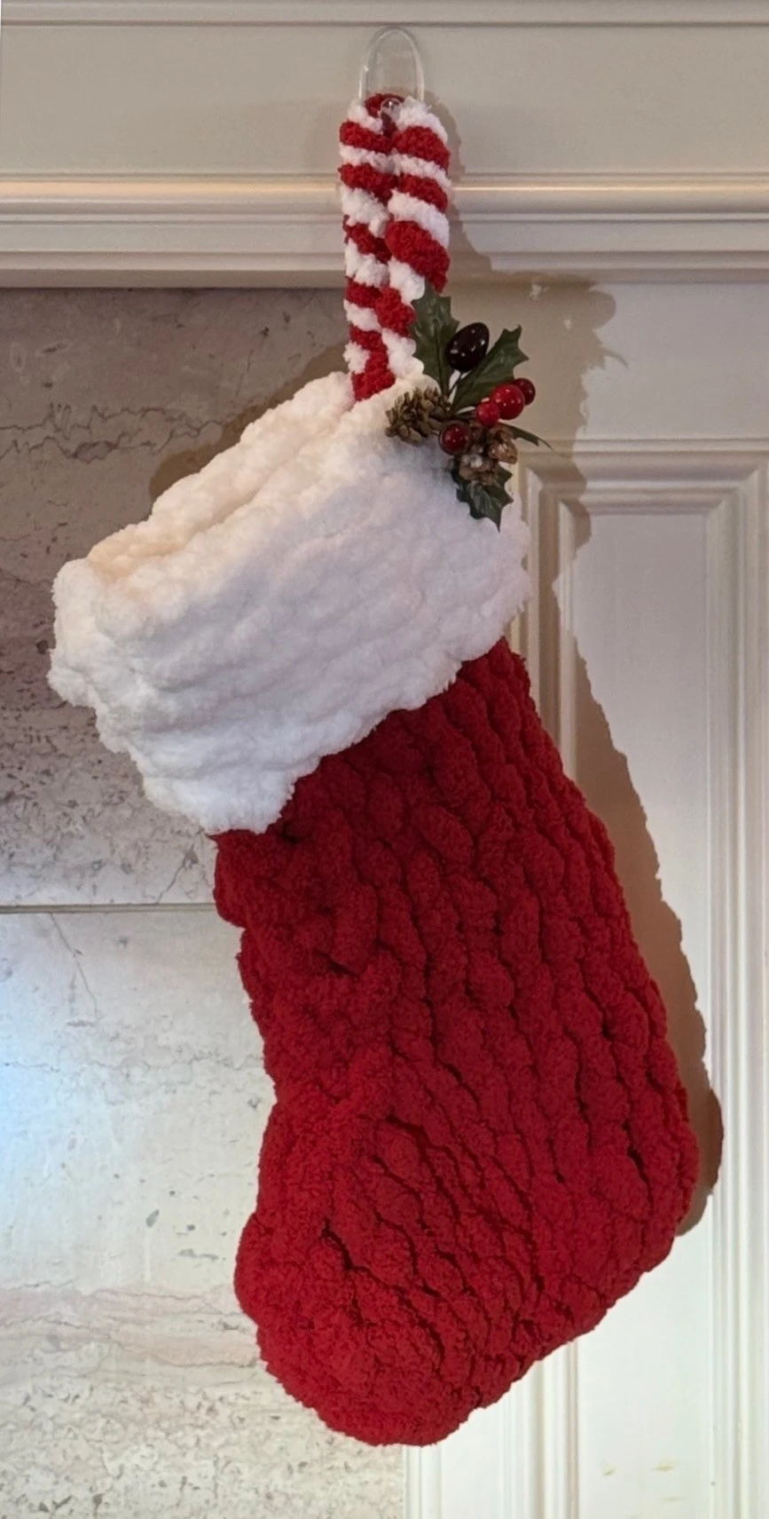 A red Christmas stocking with a white cuff and a striped hanging loop, artfully decorated with holly and berries, hangs on a white wall, showcasing the timeless charm of the art of knitting.
