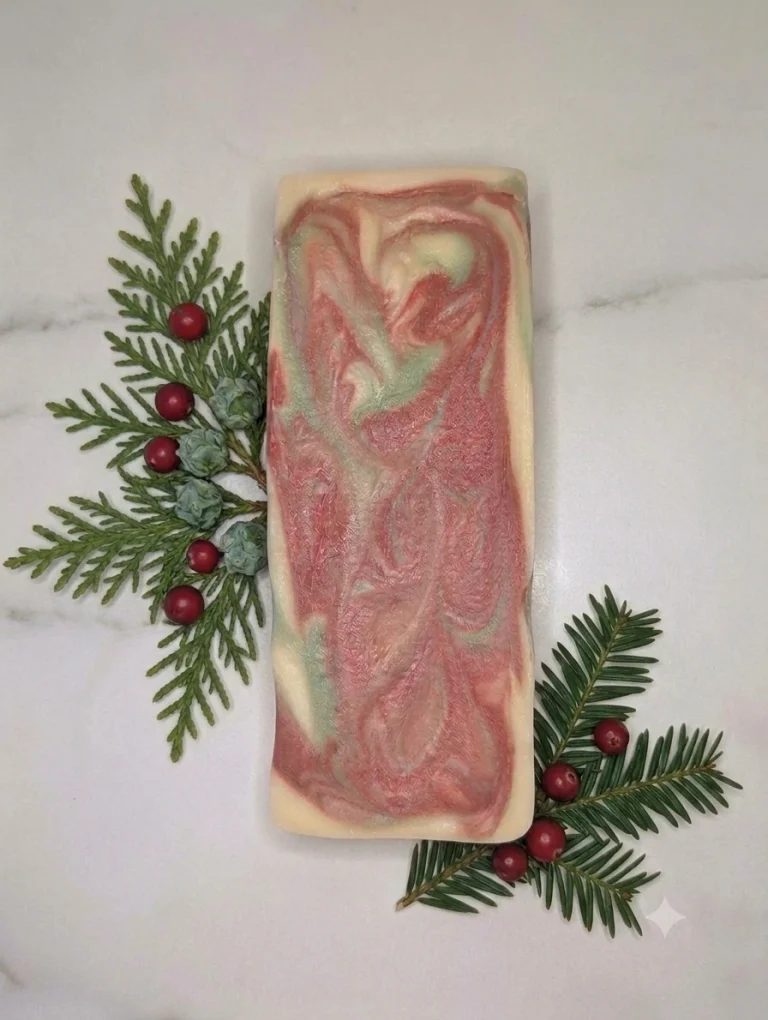 A rectangular bar of swirled pink, green, and white soap—crafted during a scented soapmaking workshop—is placed on a white surface with evergreen sprigs and red berries arranged beside it.