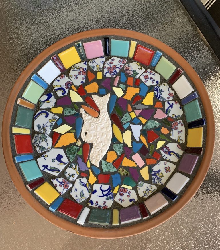 A round terra cotta tray from the Sherman Sowers Gift Faire, decorated with a mosaic of colorful tiles and floral ceramic shards surrounding a central white dove figure.