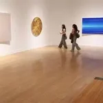 Two people walk through Laguna Art Museum’s art exhibition with wooden floors, observing abstract artworks displayed on white walls during Opening Week.