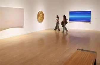 Two people walk through Laguna Art Museum’s art exhibition with wooden floors, observing abstract artworks displayed on white walls during Opening Week.