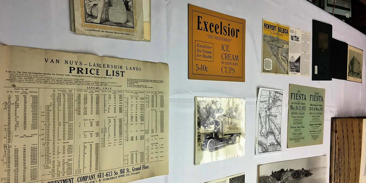 A collection of vintage advertisements, maps, and a price list for Van Nuys-Lankershim lands displayed on a white wall.